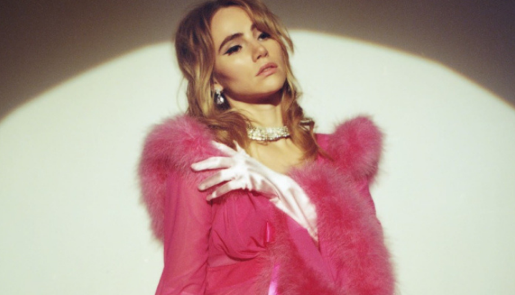 Suki Waterhouse is wearing a hot pink fur collared sheer robe. She is also wearing light pink silky gloves. Her hair is in soft curls, she is wearing a diamon choker and large dangling diamond earrings.