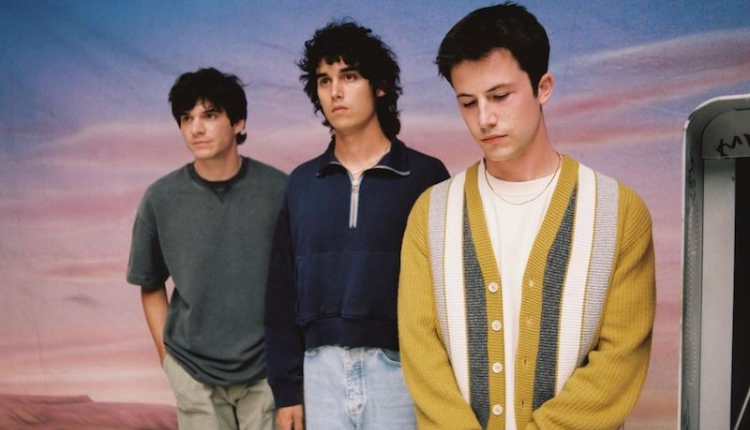 Dylan Minnette, Cole Preston, and Braeden Lemasters are standing in front of a sunset backdrop. Minnette is at the front wearing a yellow cardigan looking down at the ground. Preston is behind him wearing a navy blue jacket and light wash jeans he is looking ahead to the right. Lemasters is last wearing a green sweater and chino pants, he is looking ahead to the right as well.