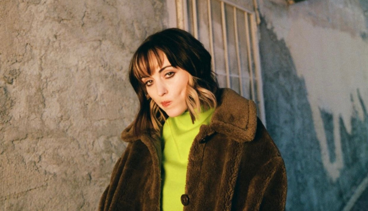 Sasha Alex Sloan is swearing a neon green shirt underneath a brown faux fur coat. her mouth is turned to the left; giving a pensive look.