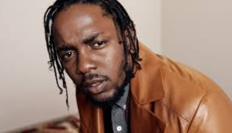 Kendrick lamar is seen wearing awarm mustard colored jacket with a gray collared shirt underneath and looking directly into the camera