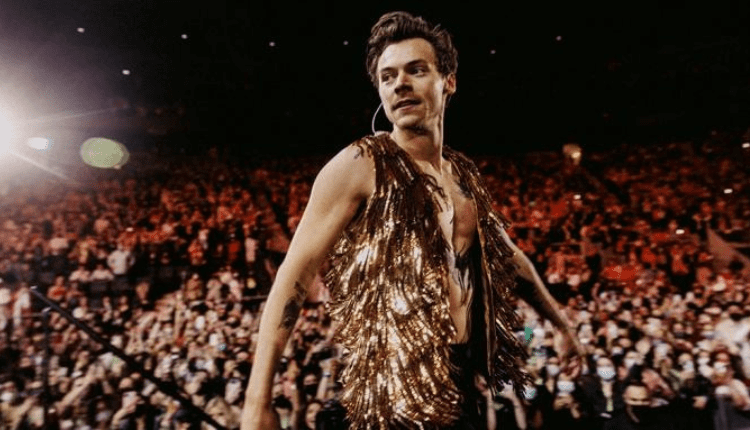 Harry styles is seen wearing a gold fringe vest exposing his bare chest, he is performing in front of a large audience