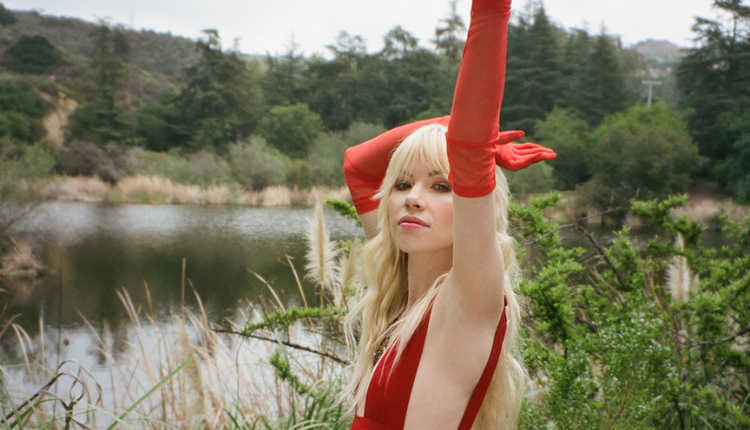 Carly Rae Jepsen is wearing red gloves and a red dress out by a lake, she is posing eleganty with her hands up