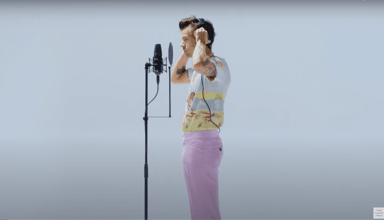Harry styles adjusts his headphones as he prepares to sing on first take he is wearing pastel pink oants and a colrful shirt