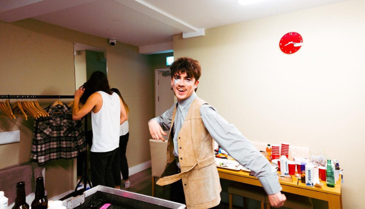 declan mckenna is seen backstage happily looking at the camera, he is wearing his famous stage make up and a beige vest over a light blue long sleeve button up