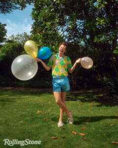 Harry Styles in a field cheerfully holding four balloons. He's wearing a yellow, white, and green beaded polo top, with sparkly blue shorts.