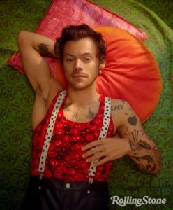Harry Styles laying down in a cherry print tank top, black pants, and polka dot suspenders.