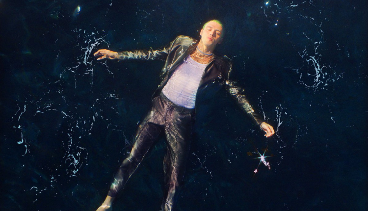 Lauv is floating in a alrge body of water while wearing a long pants, sports coat, and white tnak top