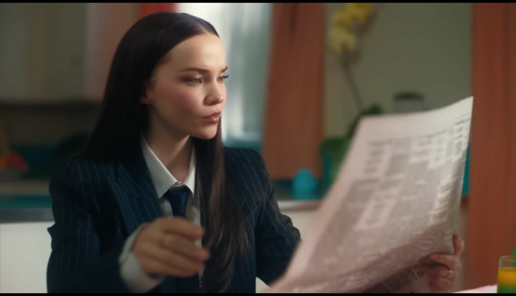 Dove Cameron is seen wearing a black business suit and reading the newspaper, a still from her music video "breakfast"