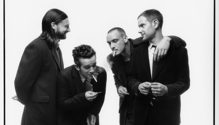 the 1975 from L to R (Ross Mcdonald, Matty Healy, George Daniels, Adam Hann) are huddled around each other, wearing posh suits, Healy and Daniels are seen smoking, Mcdonald and Hann are seen smiling