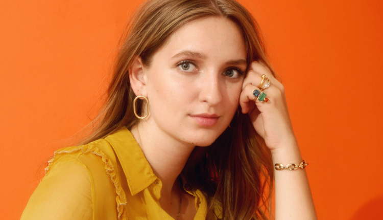 Madison Cunningham is seen wearing a yellow collared shirt, gold hoop earrings, and jewelry decorating her hands and wrist. She is in front of an orange background