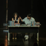 Singer-songwriter BIBI sitting at a desk full of books, performing a skit on stage with one of her dancers.