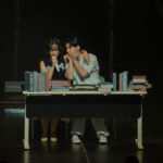 Singer-songwriter BIBI sitting at a desk full of books, performing a skit on stage with one of her dancers.