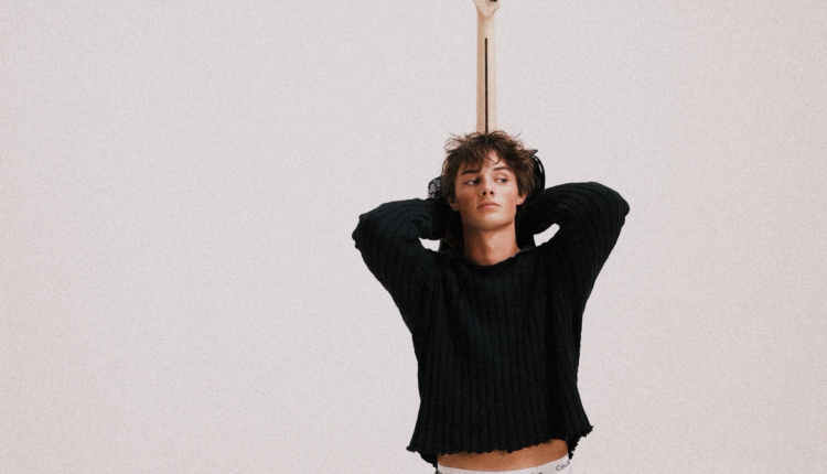 Thomas is wearing a black sweater, standing against a white wall, and holding a guitar behind his back.